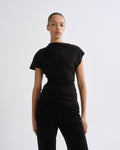 A model wearing a black short-sleeve top with a draped, asymmetrical neckline and black pants, photographed against a plain light gray studio background.