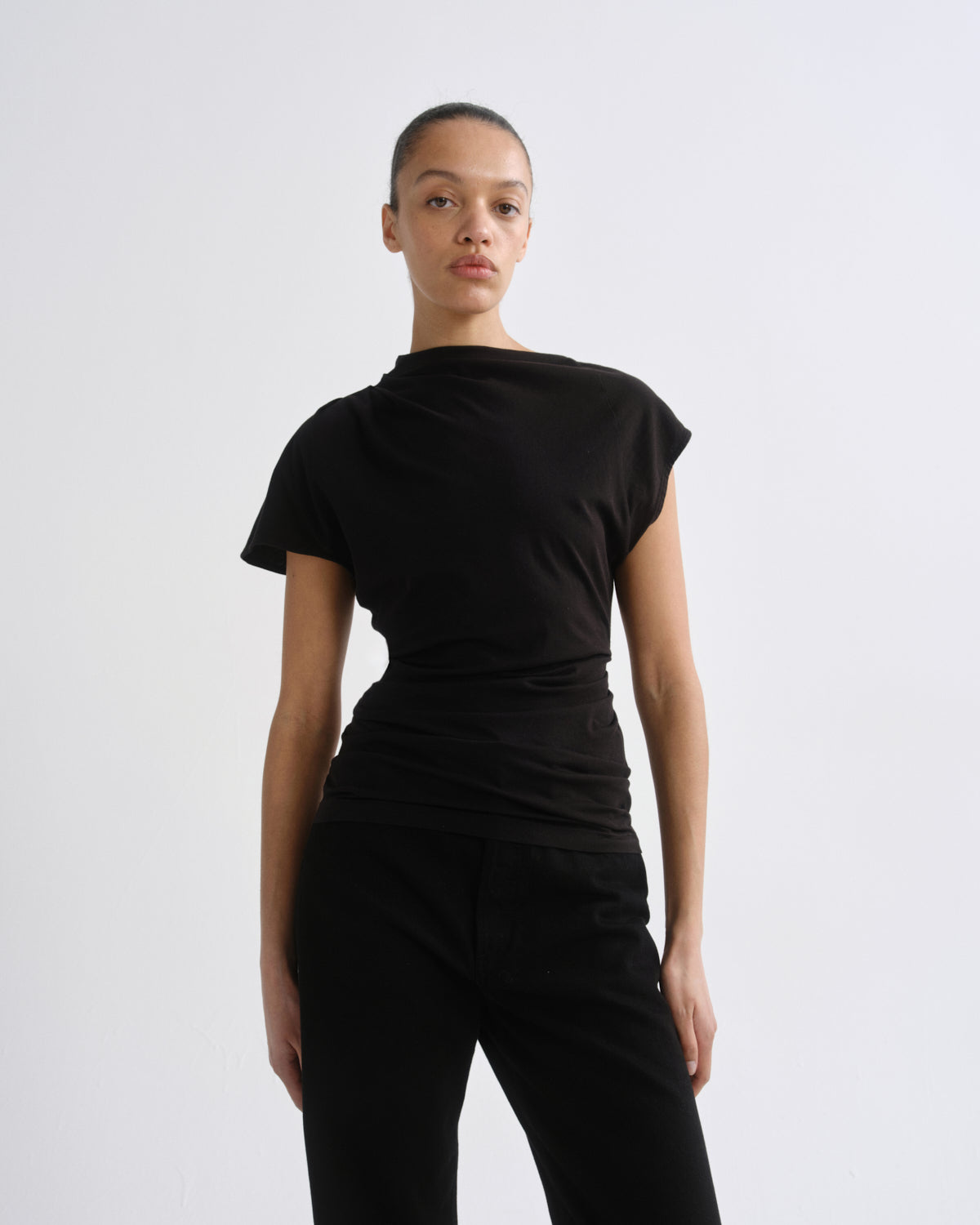 A model wearing a black short-sleeve top with a draped, asymmetrical neckline and black pants, photographed against a plain light gray studio background.