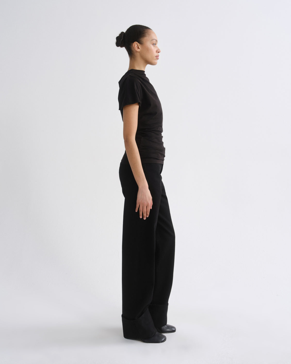 A model in a side profile wearing a black short-sleeve mock neck top and matching black wide-leg trousers, standing against a light gray studio background.