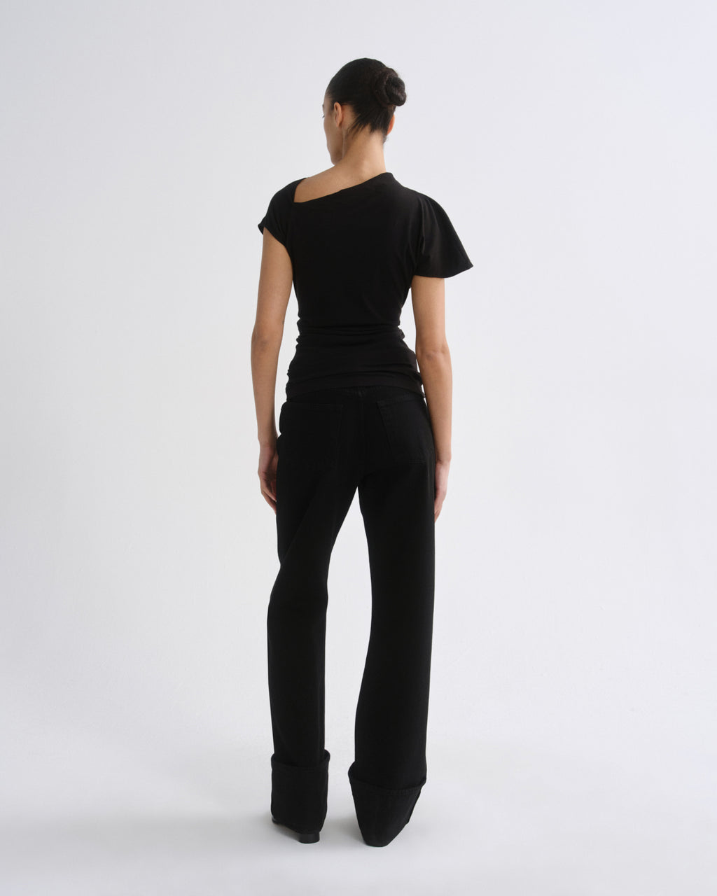 A woman viewed from the back wearing a black asymmetrical short-sleeve top and black wide-leg pants against a plain light gray studio background.