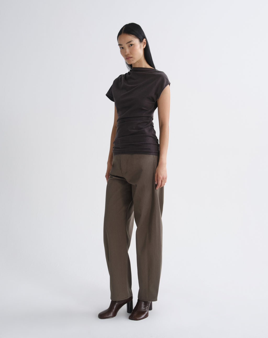 Person wearing a dark brown draped cap sleeve top with olive wide leg pants viewed from the side on a grey background