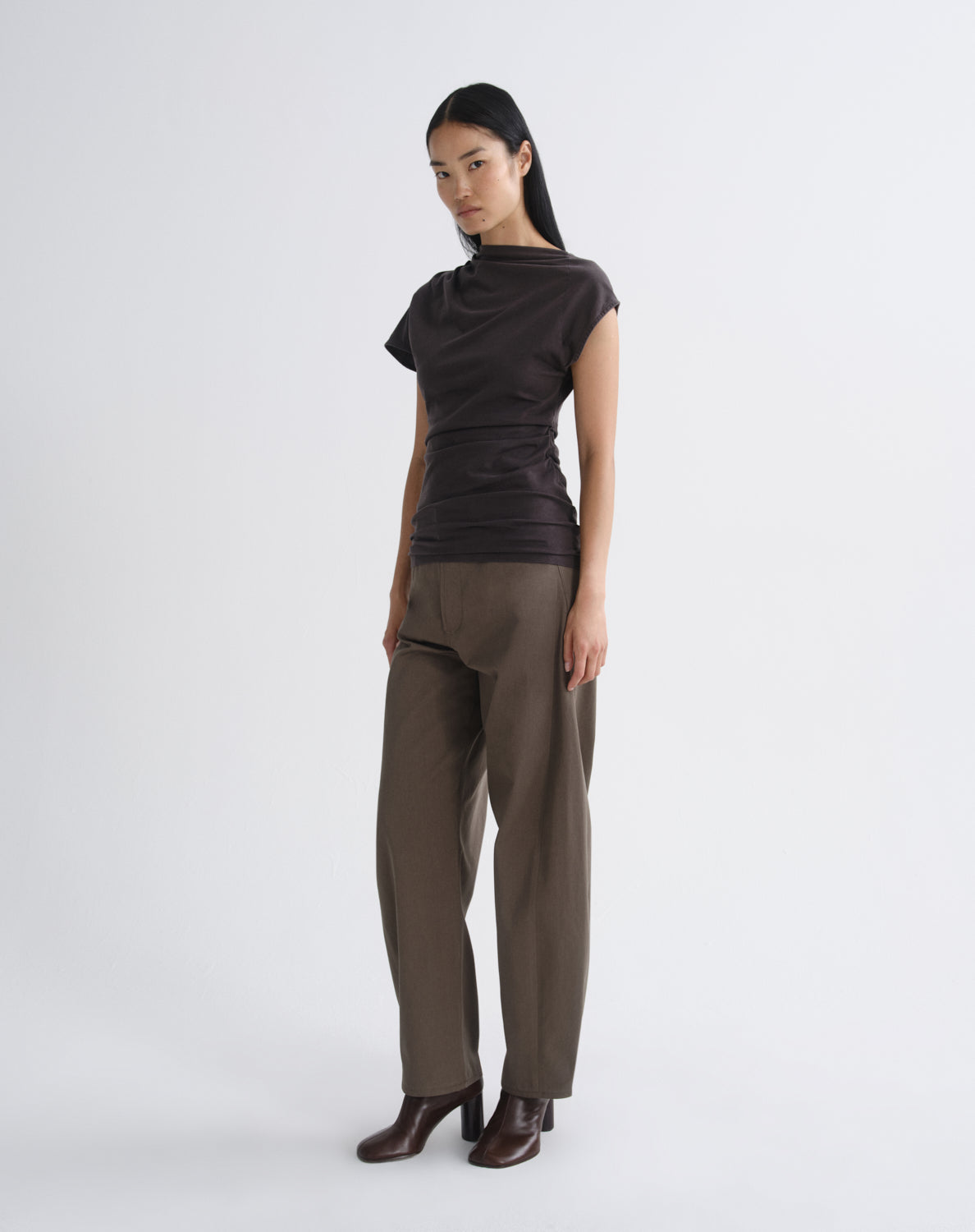 Person wearing a dark brown draped cap sleeve top with olive wide leg pants viewed from the side on a grey background