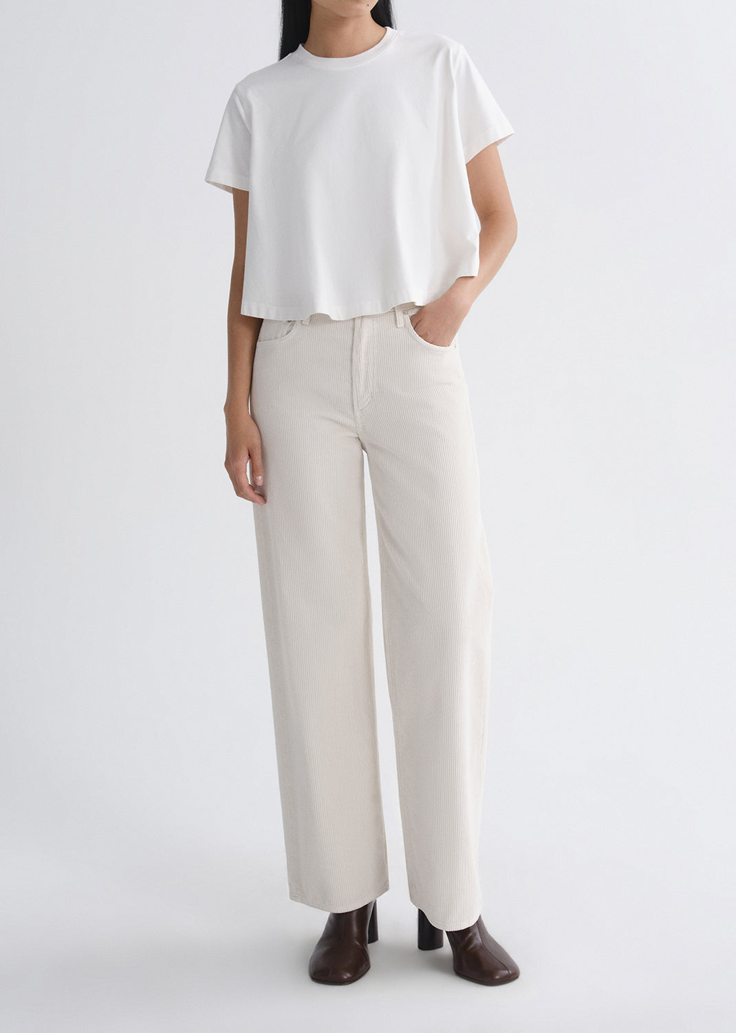 A model wearing a white short-sleeve cropped t-shirt and off-white wide-leg trousers, paired with dark brown heeled shoes, against a grey background.