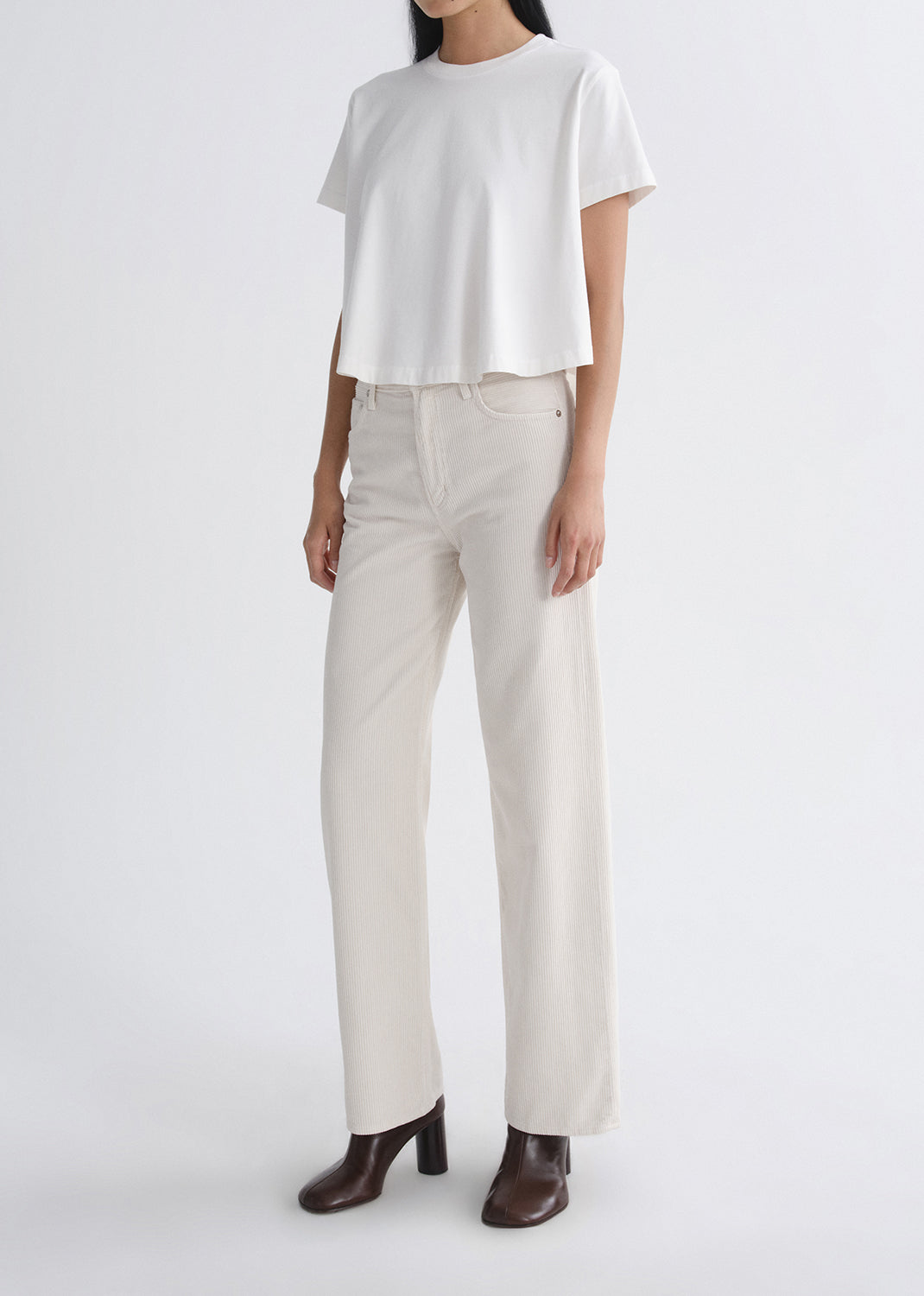 A model wearing a white short-sleeve cropped t-shirt, off-white wide-leg trousers, and dark brown heeled ankle boots, photographed against a grey background.