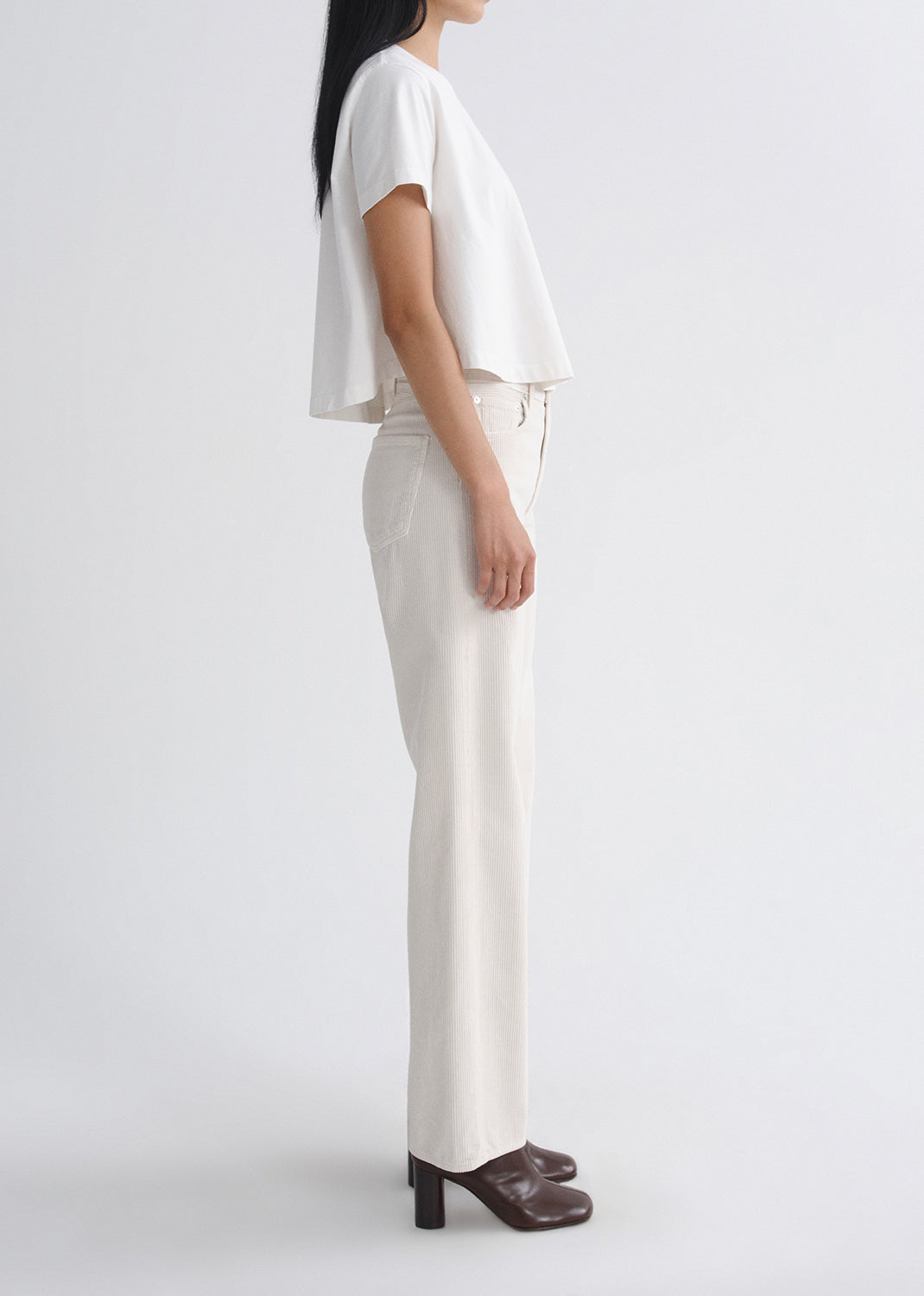 A model shown from the side wearing a white short-sleeve cropped top and off-white straight-leg trousers, paired with dark brown mule heels, against a grey background.