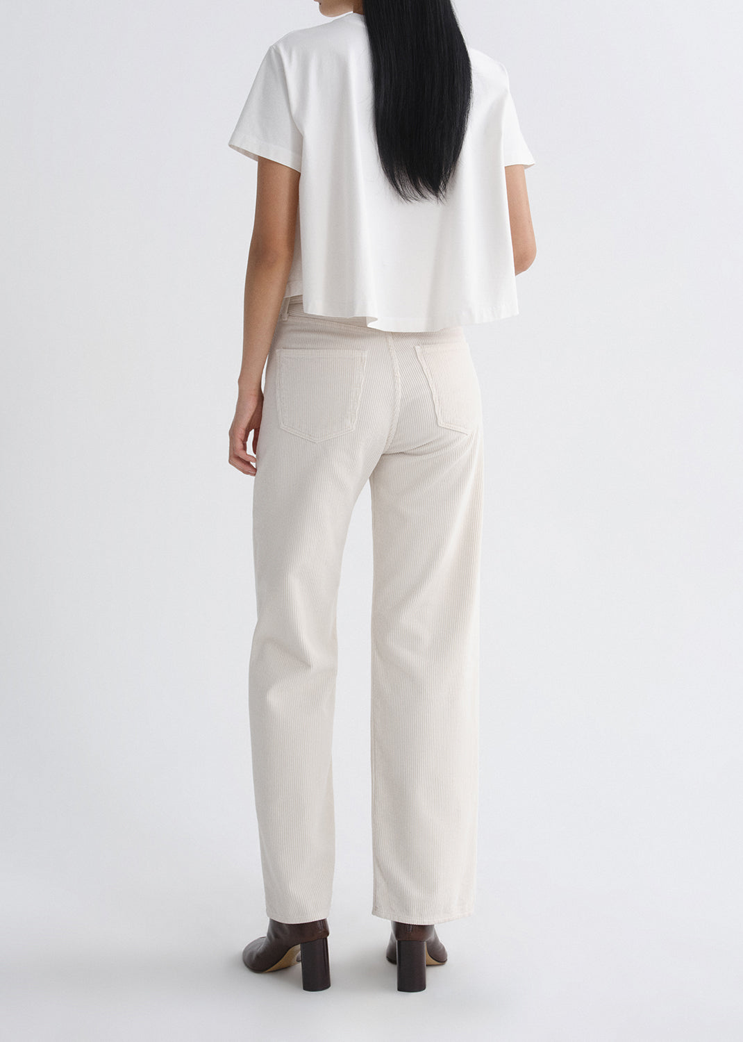 A woman shown from the back wearing a white short-sleeve cropped top and off-white straight-leg trousers, paired with dark brown heeled shoes, against a grey background.