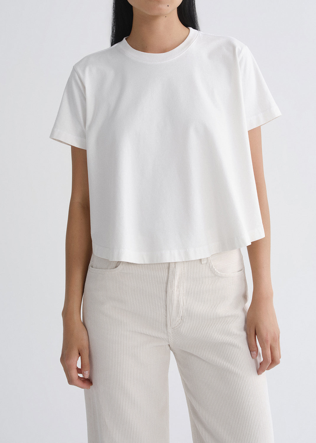 A model wearing a white short-sleeve cropped t-shirt and off-white corduroy trousers, shown from the neck down against a grey background.