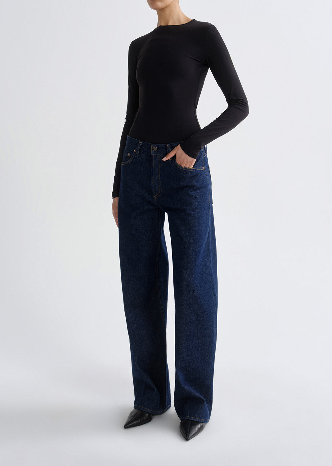 A model wearing a black long-sleeve mock-neck top and dark blue wide-leg jeans, standing against a grey background.