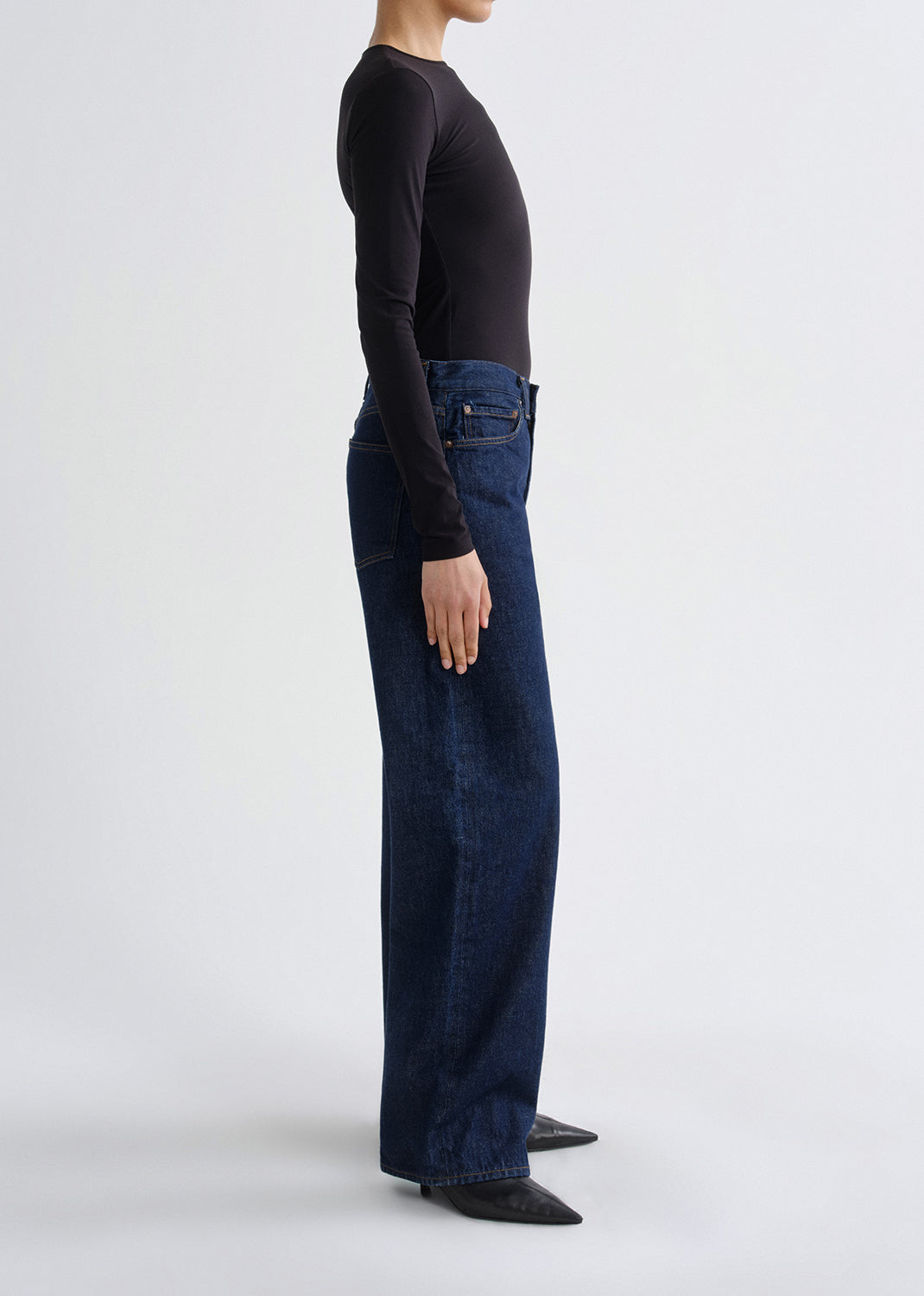 A side-profile studio shot of a model wearing dark blue wide-leg jeans, a black long-sleeve top, and black pointed shoes against a grey background.