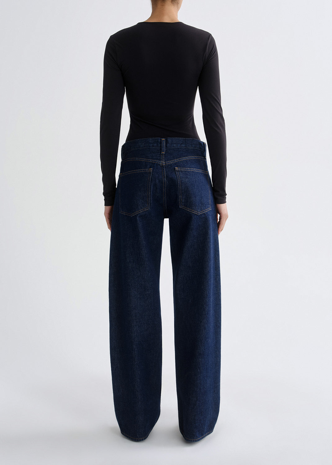 A model shown from the back wearing a black long-sleeve top and dark blue wide-leg denim jeans against a grey background.