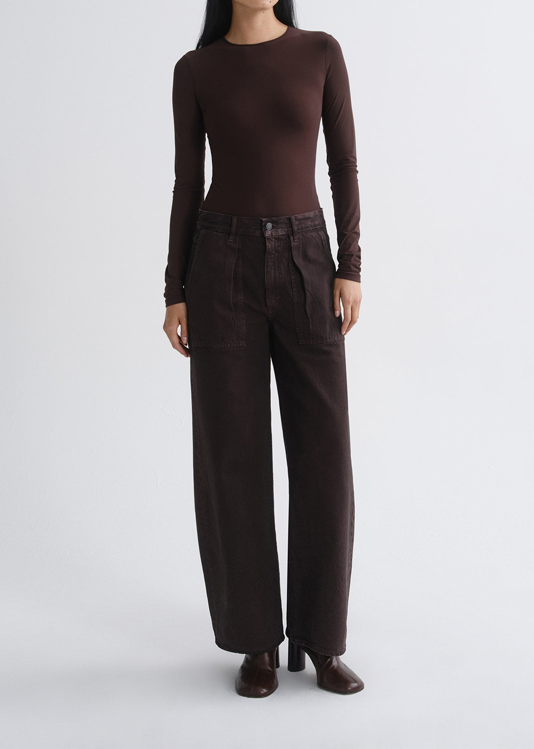 A model wearing a dark brown long-sleeve crewneck top and matching dark brown wide-leg trousers, standing against a grey background.