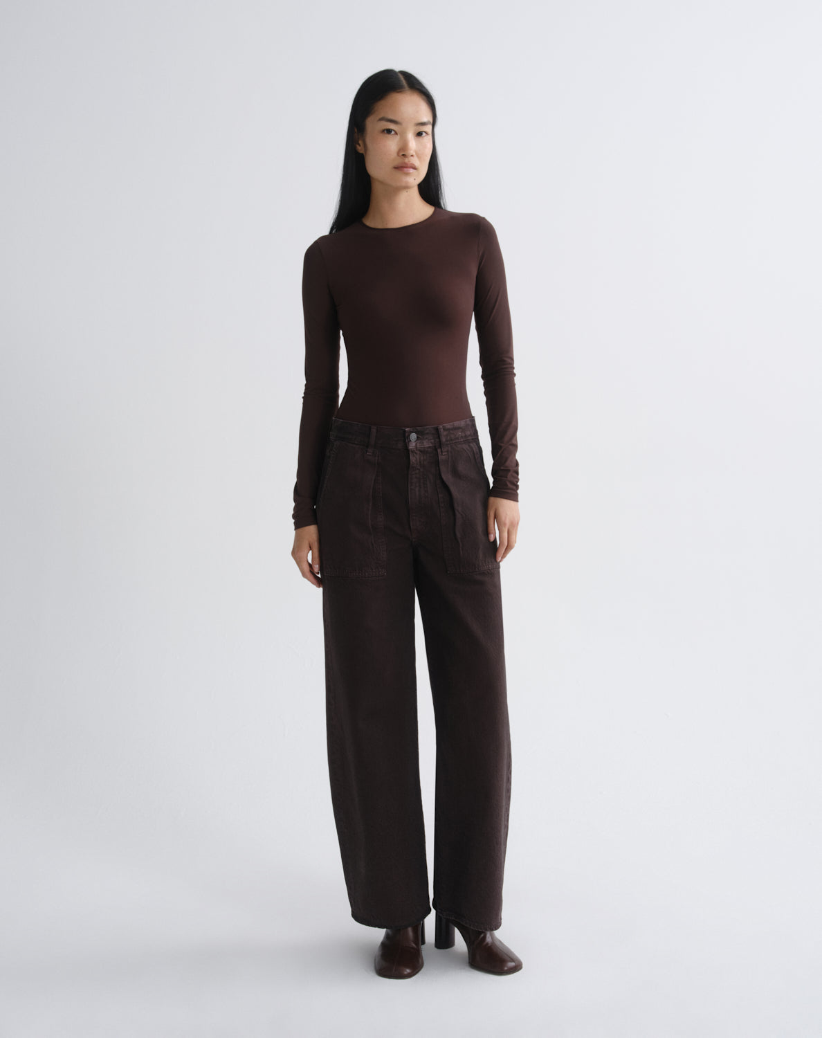 A model stands in a studio wearing a dark brown long-sleeve crewneck top and matching dark brown wide-leg trousers, paired with dark brown shoes, against a light gray background.