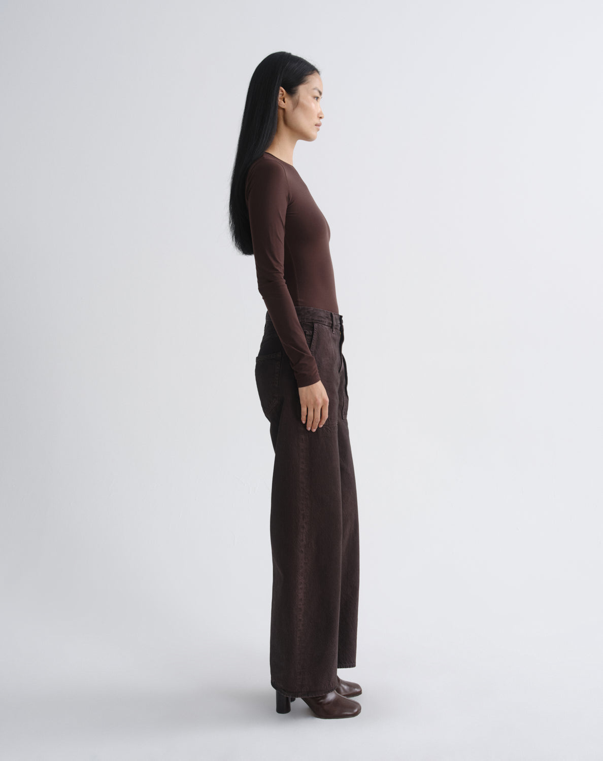A woman in a side profile wearing a dark brown long-sleeve top and matching wide-leg trousers, standing against a light gray studio background.