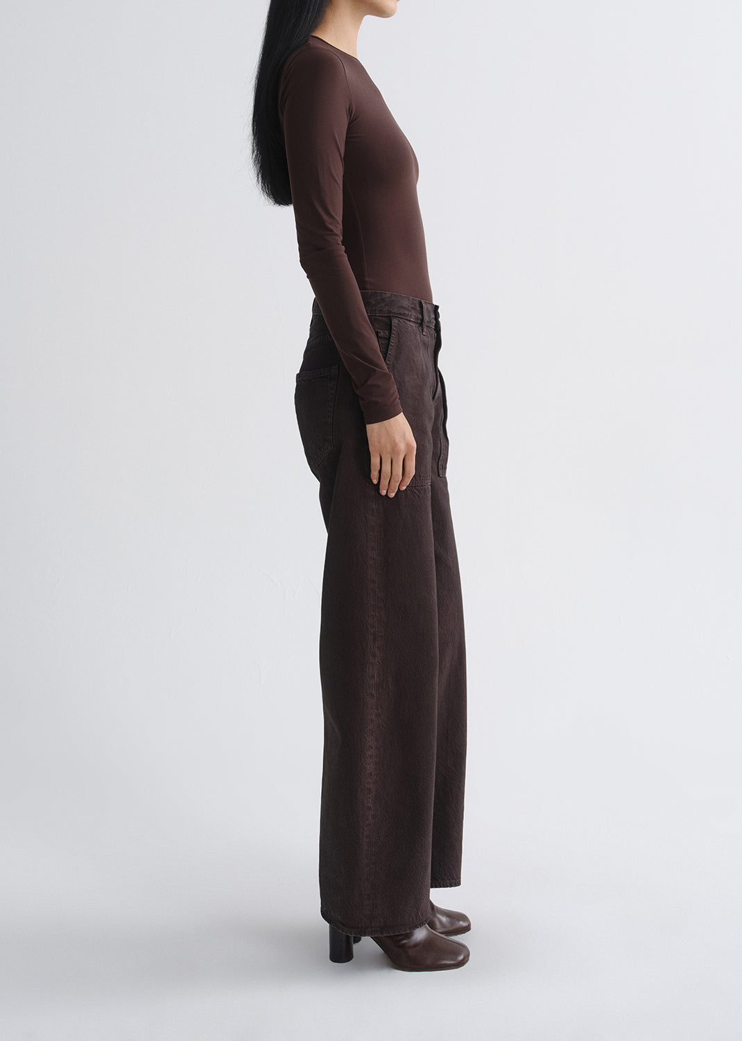 A model shown in side profile wearing a dark brown long-sleeve top and matching dark brown wide-leg trousers against a grey background.