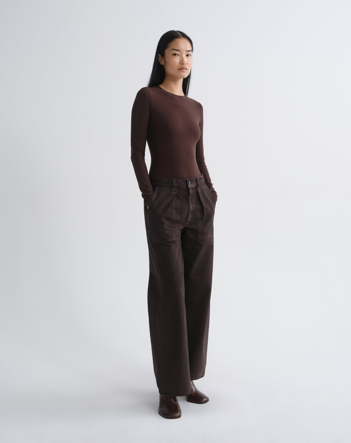 A model stands in a studio wearing a dark brown long-sleeve crewneck top and matching dark brown wide-leg trousers, paired with dark brown shoes, against a light gray background.