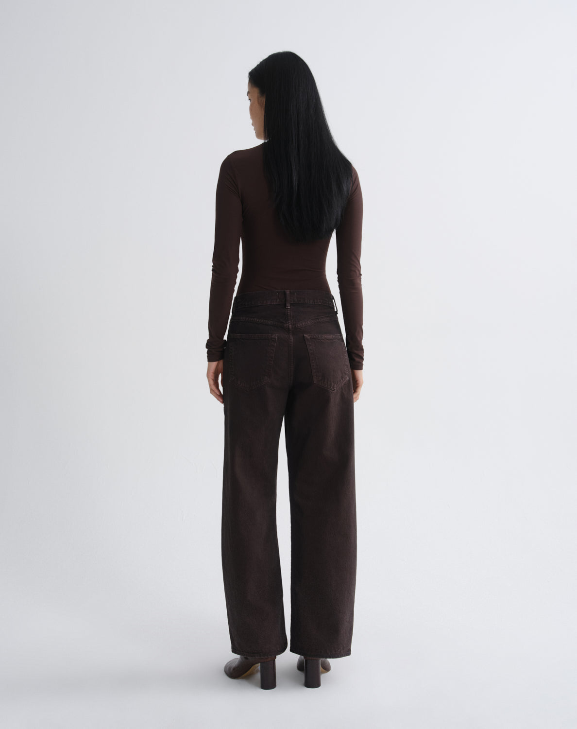 A woman with long black hair is shown from the back wearing a dark brown long-sleeve top and matching dark brown wide-leg trousers, standing against a light gray studio background.