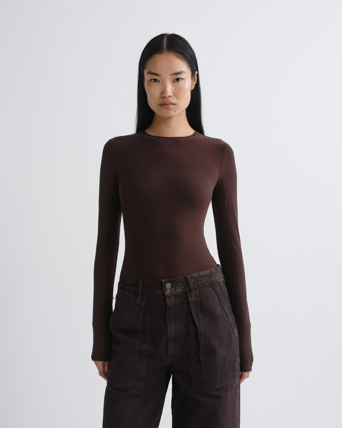 A model wearing a dark brown long-sleeve crewneck top and dark brown high-waisted pants, standing against a light gray studio background.