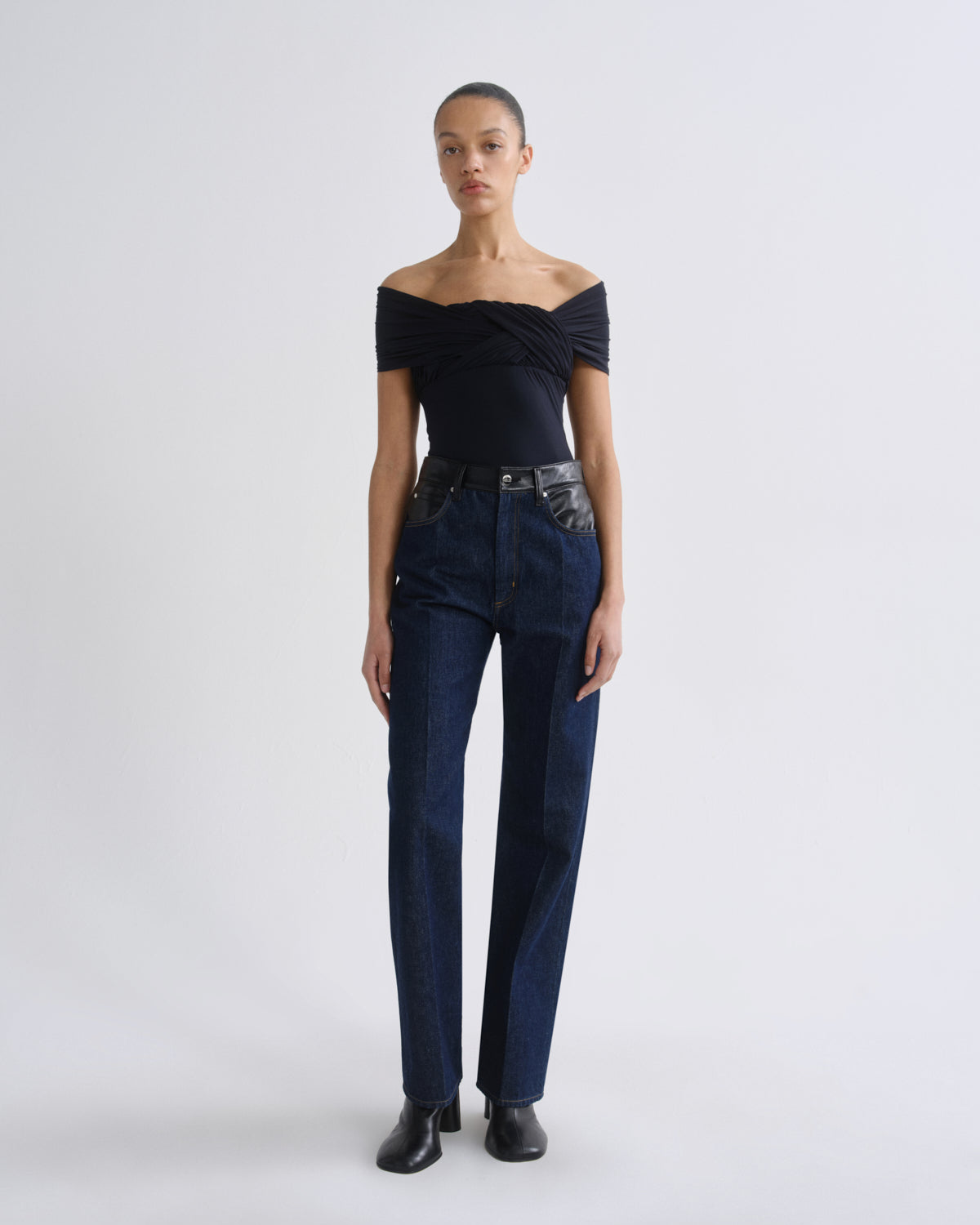 A model stands against a light gray studio background wearing a dark off-the-shoulder top, dark blue straight-leg jeans, and black shoes.