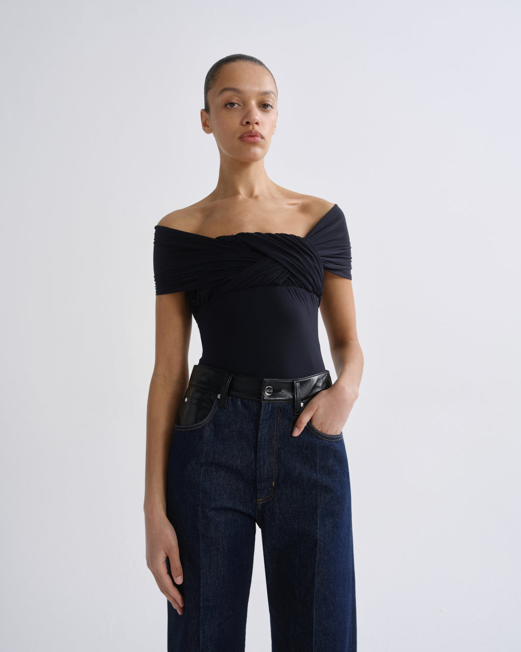 A model wearing a black off-the-shoulder bodysuit with a twisted front detail, paired with dark blue jeans and a black belt, standing against a light gray studio background.