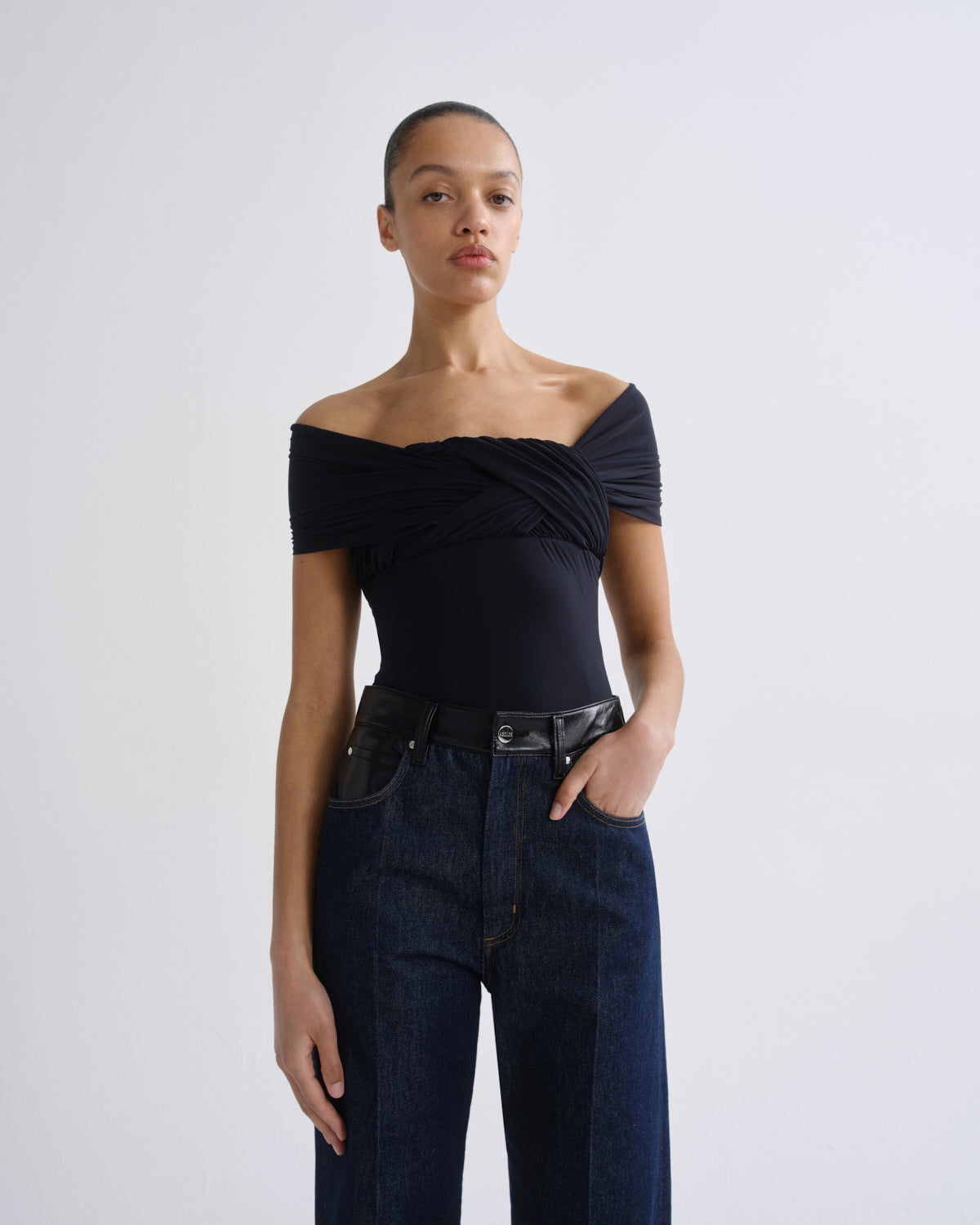 A model wearing a black off-the-shoulder bodysuit with a twisted front detail, paired with dark blue jeans and a black belt, standing against a light gray studio background.