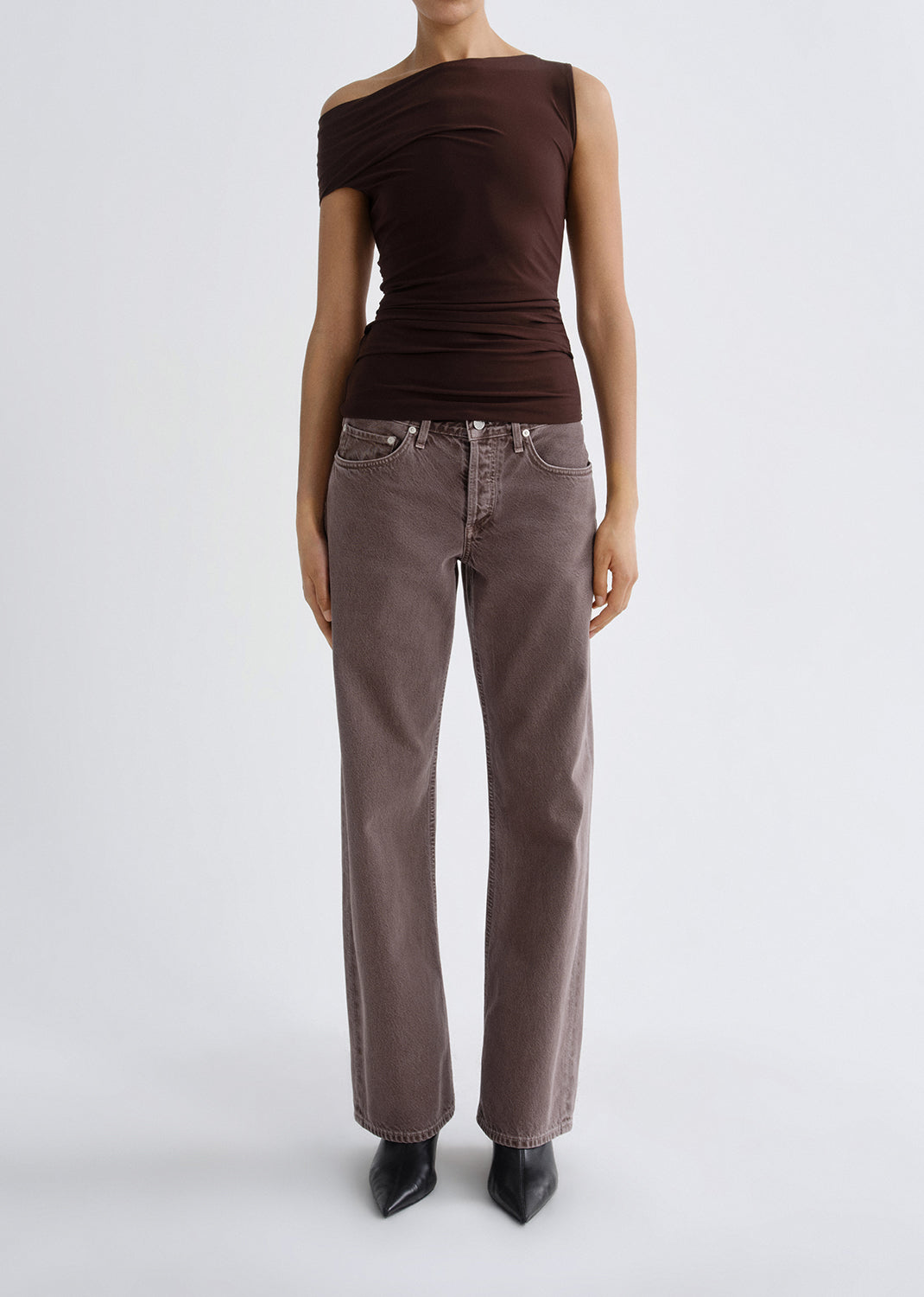 Person wearing a dark brown one shoulder top with mauve straight leg jeans on a grey background.