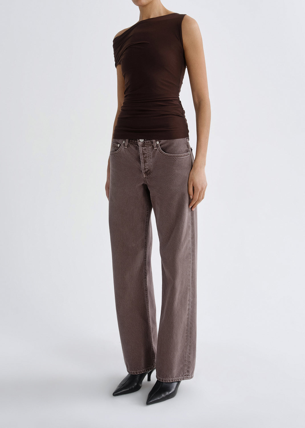 Person wearing a dark brown one shoulder top with mauve straight leg jeans on a grey background.