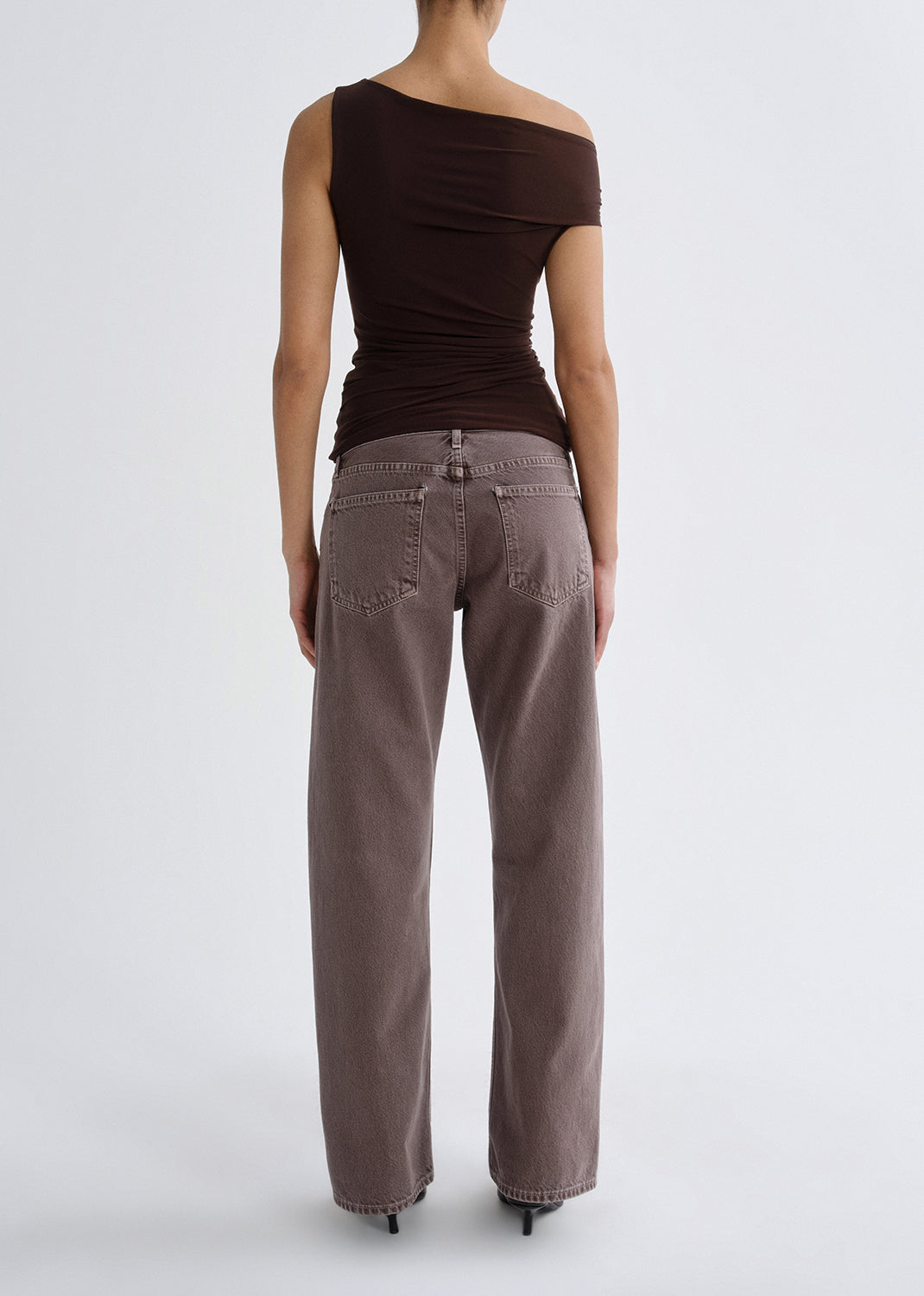 Person wearing a dark brown one shoulder top with mauve straight leg jeans viewed from the back on a grey background.