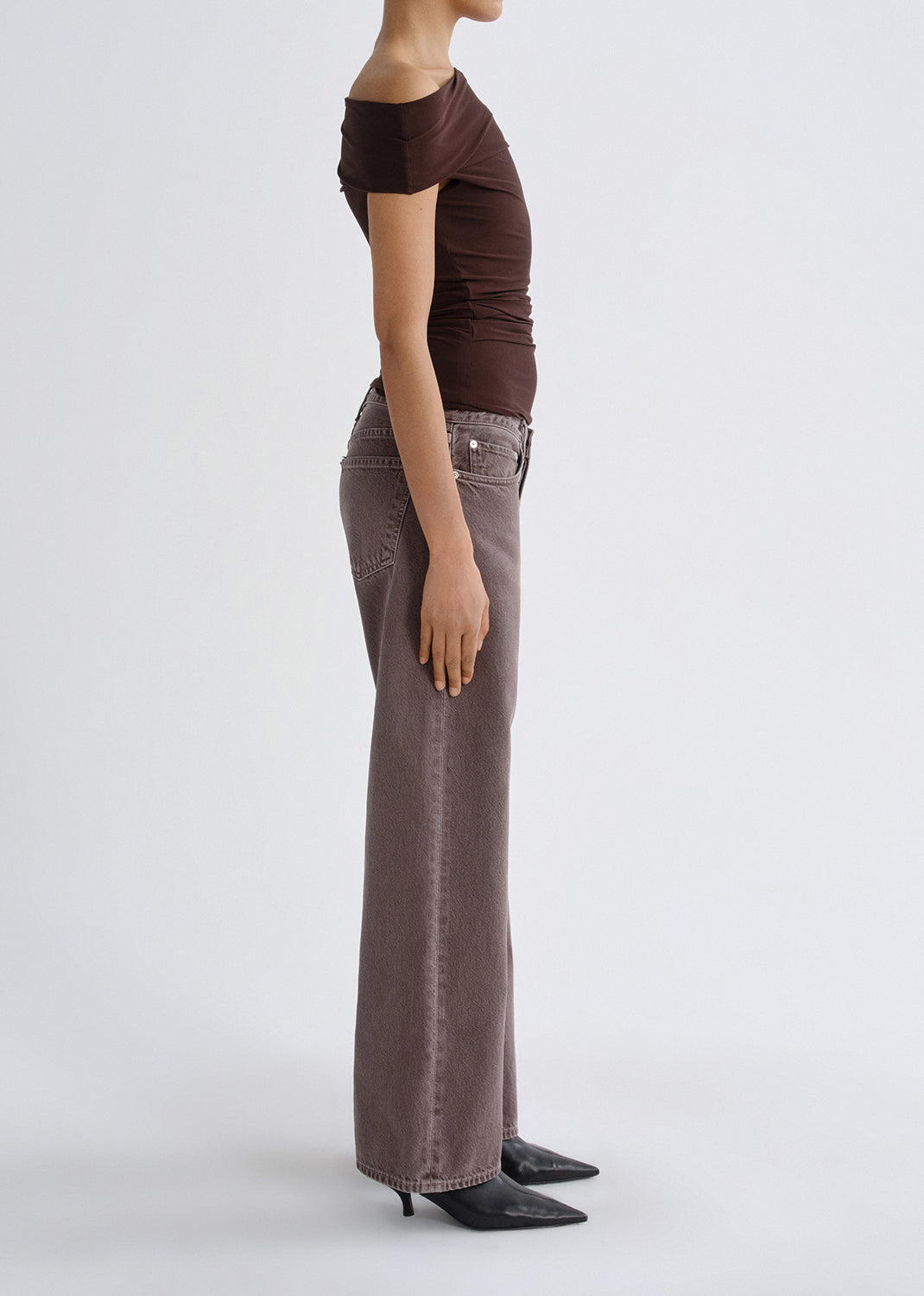 Person wearing a dark brown one shoulder top with mauve straight leg jeans viewed from the side on a grey background.