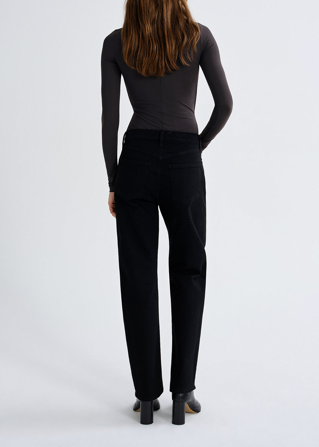 Person wearing a charcoal long sleeve ruched mock neck top with black skinny jeans viewed from the back on a grey background.