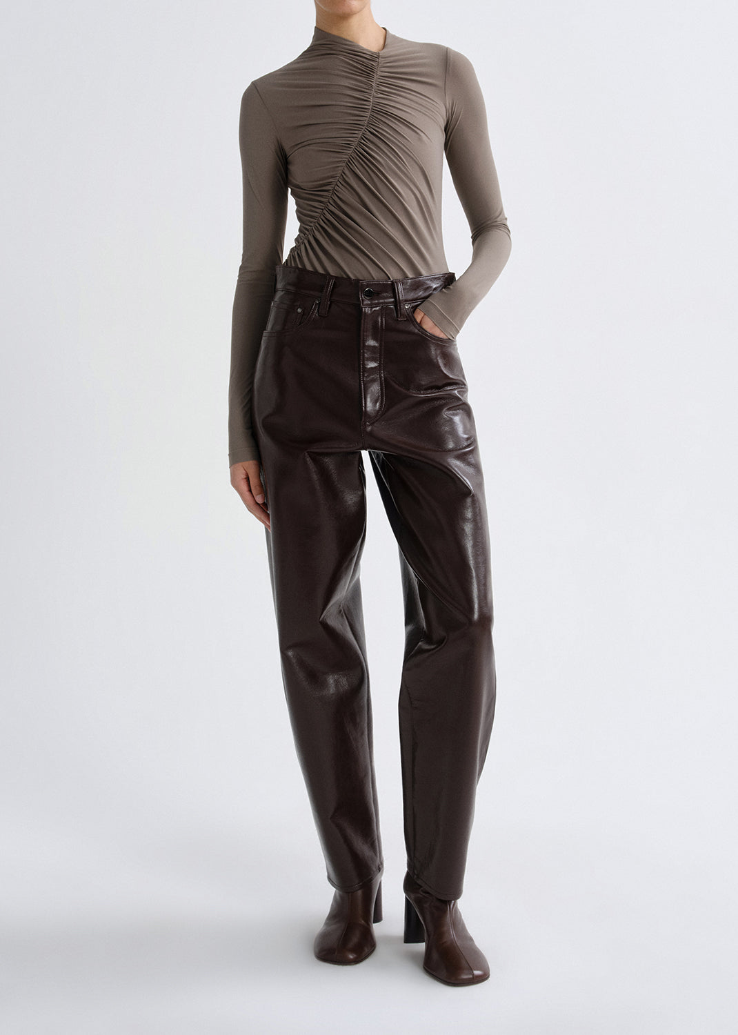 Person wearing a taupe long sleeve ruched mock neck top with dark brown leather pants on a grey background.
