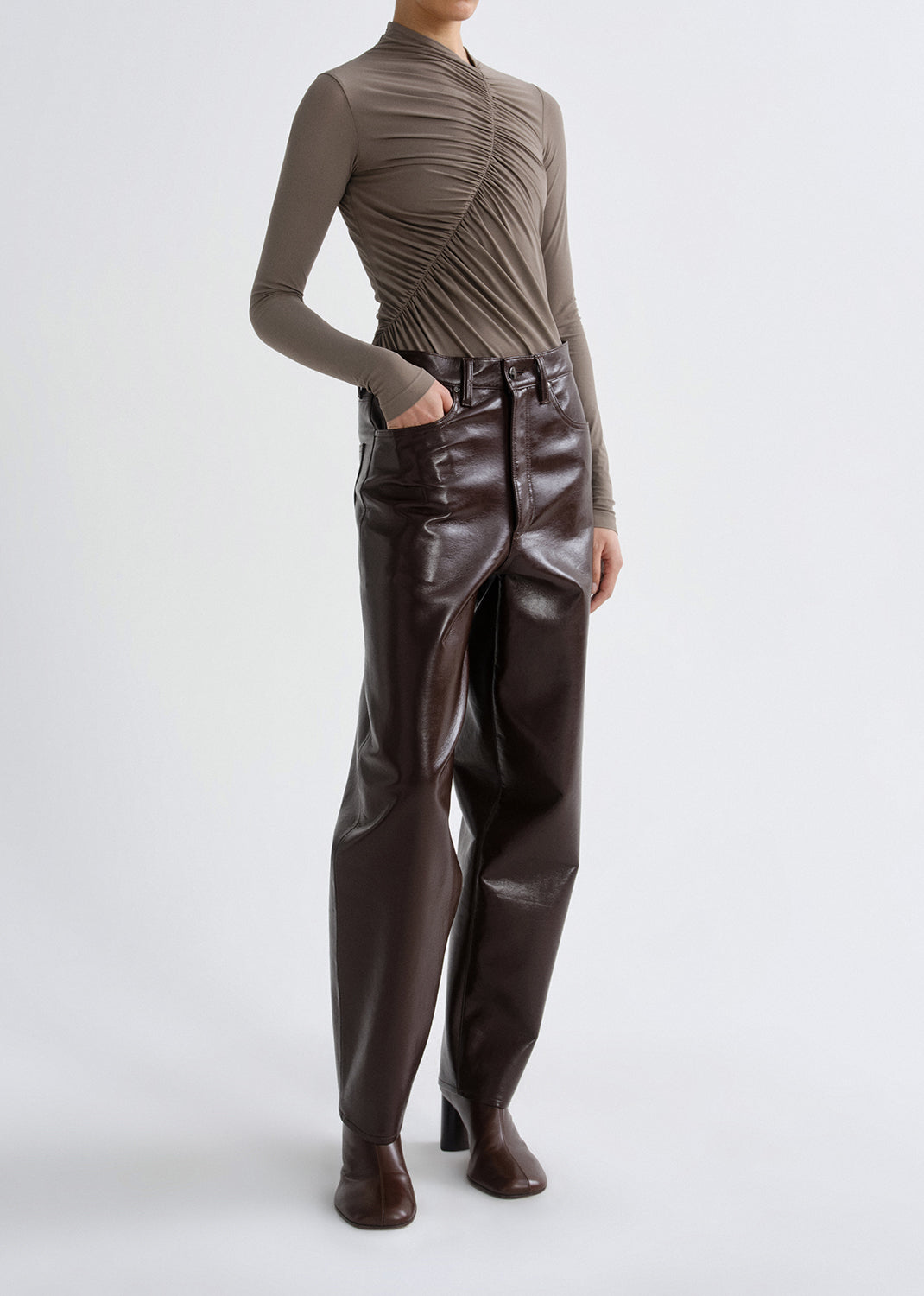 Person wearing a taupe long sleeve ruched mock neck top with dark brown leather pants on a grey background.