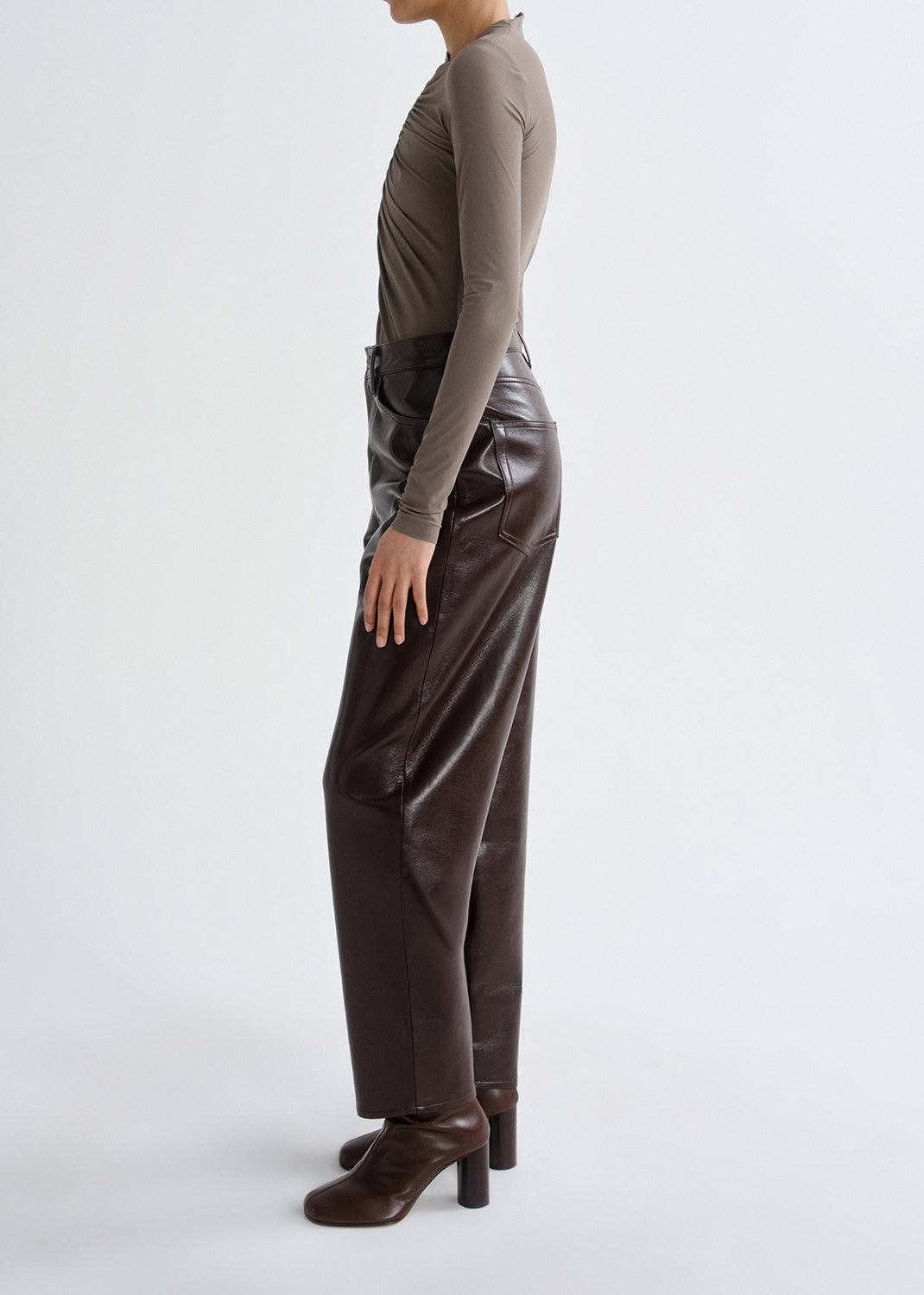 Person wearing a taupe long sleeve ruched mock neck top with dark brown leather pants viewed from the side on a grey background.