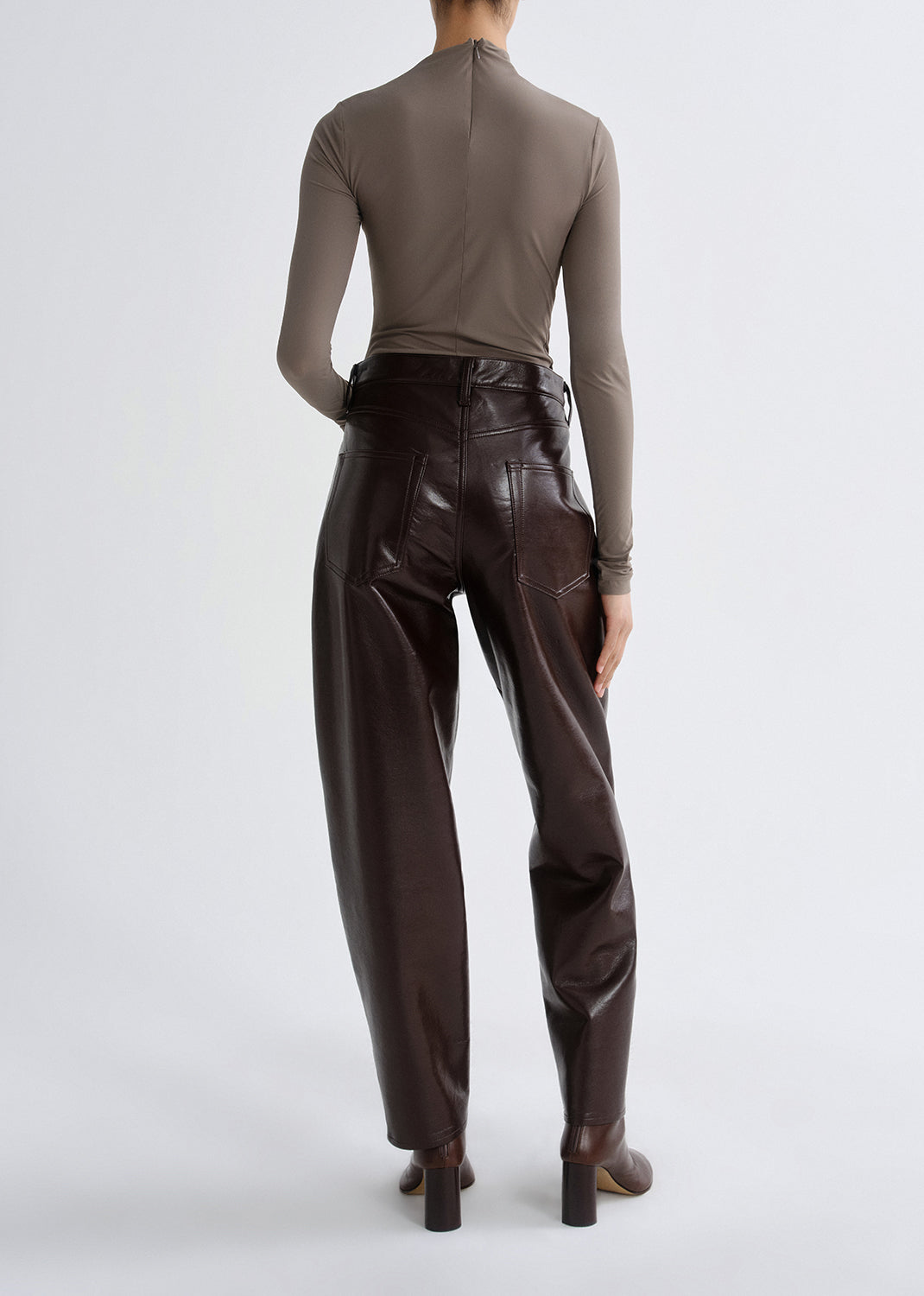Person wearing a taupe long sleeve ruched mock neck top with dark brown leather pants viewed from the back on a grey background.