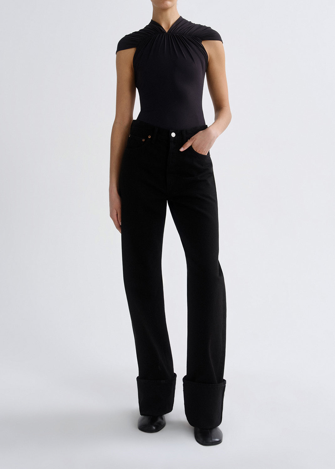 A model wearing a black sleeveless top with a gathered halter-style neckline and black wide-leg trousers with rolled cuffs, photographed against a grey background.