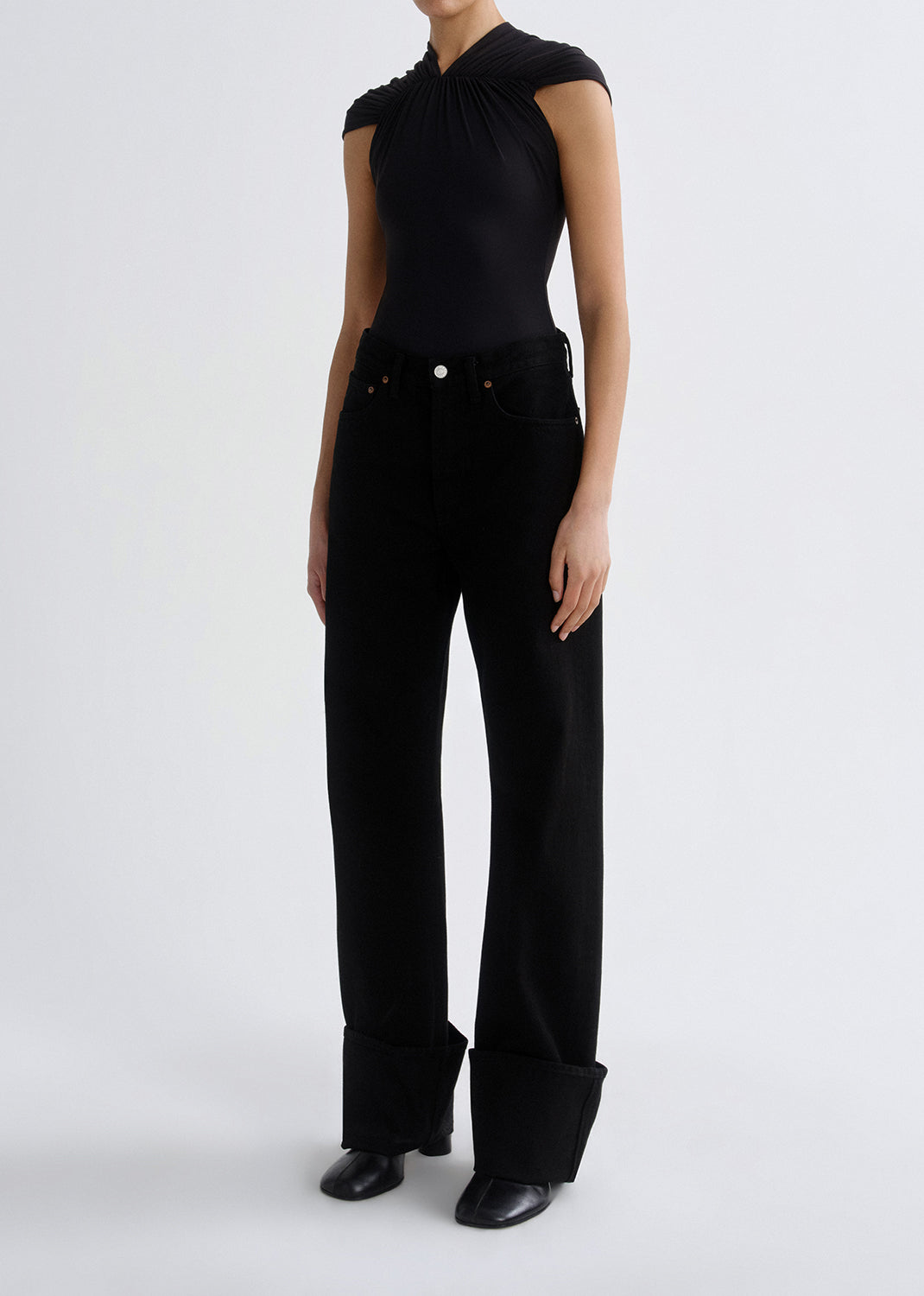 A model wearing a black sleeveless top with a gathered neckline and black wide-leg jeans with rolled cuffs, paired with black shoes, against a grey background.