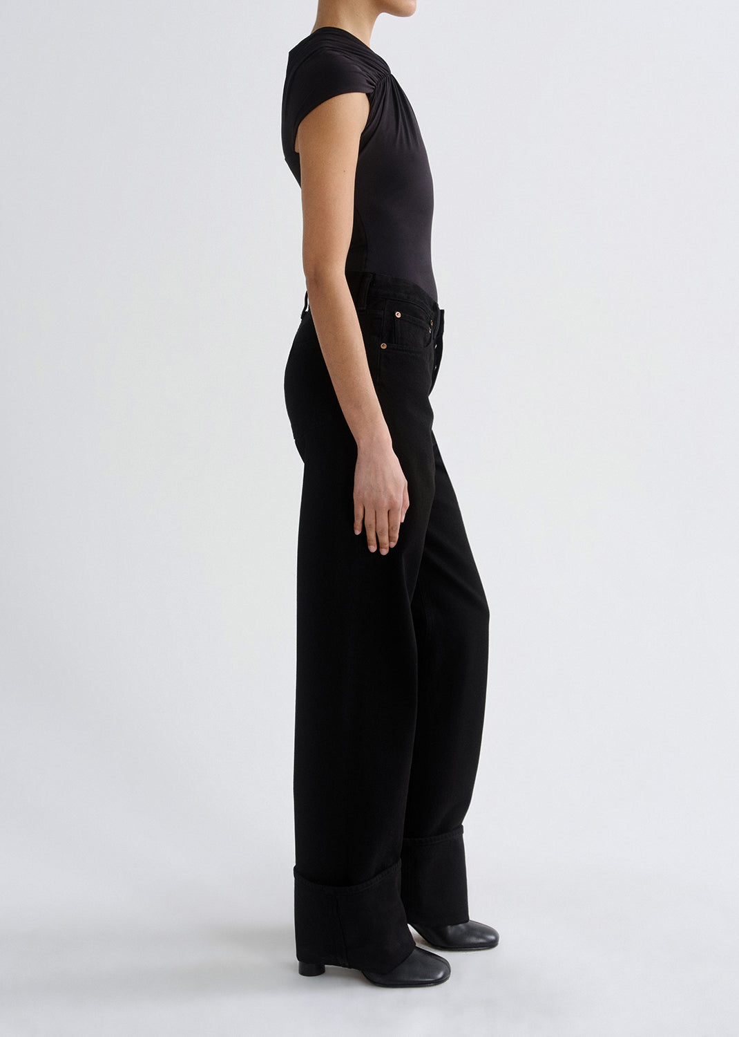 A model shown from the side wearing a black short-sleeve top and black wide-leg trousers with rolled cuffs, photographed against a grey background.