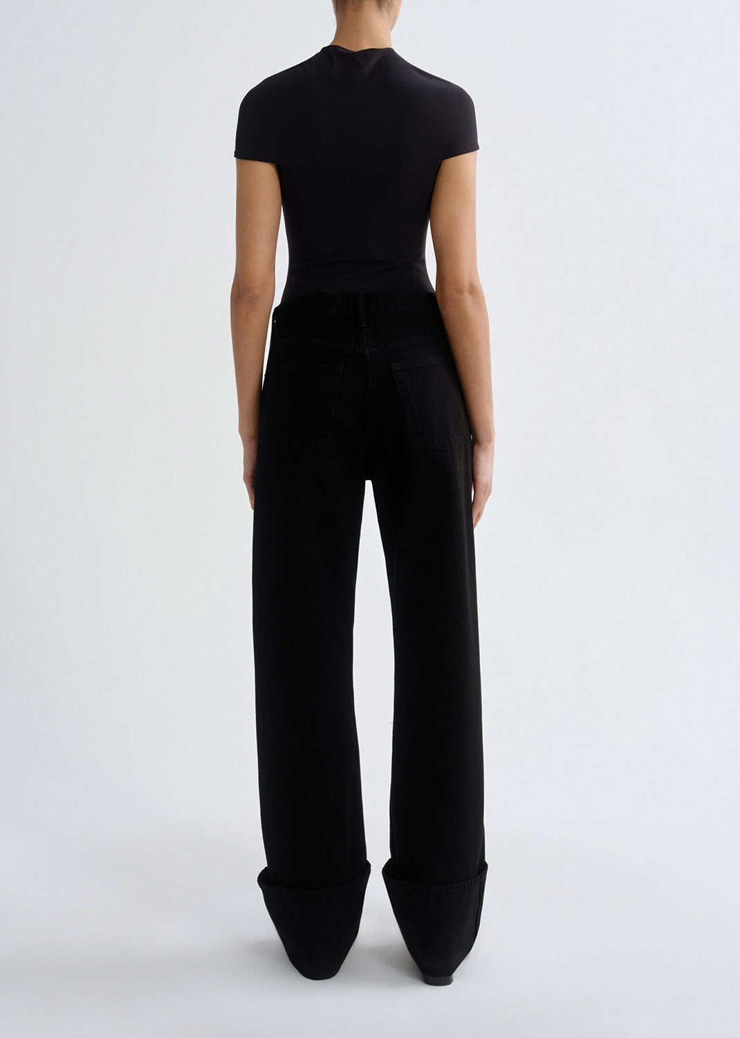 A model shown from the back wearing a black short-sleeve mock-neck top and black wide-leg trousers with pointed toes, against a grey background.