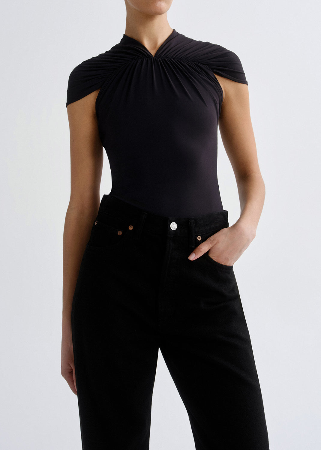 A model wearing a black sleeveless top with a dramatic gathered, draped neckline and black high-waisted jeans, shown from the neck down against a grey background.