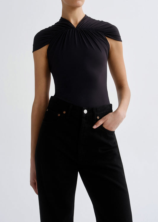 A model wearing a black sleeveless top with a dramatic gathered, draped neckline and black high-waisted jeans, shown from the neck down against a grey background.