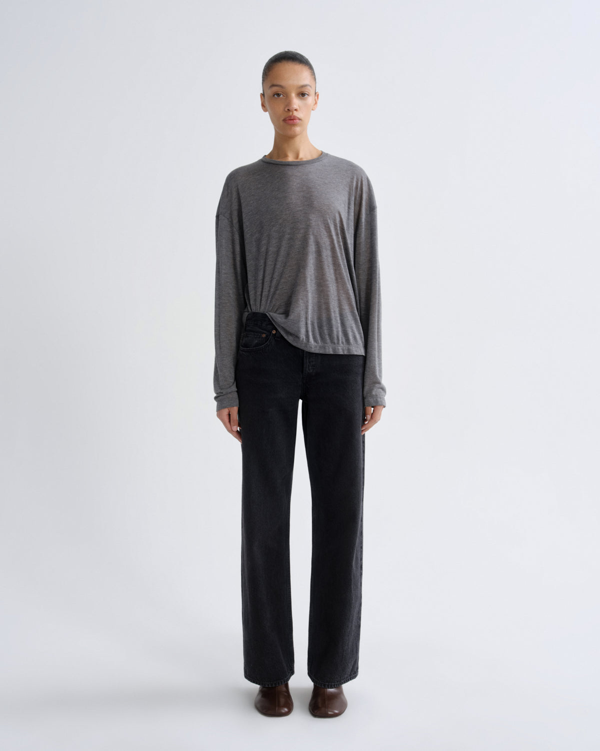 A model stands against a light gray studio background wearing a heather gray long-sleeve knit top tied at the waist with black wide-leg trousers and brown shoes.