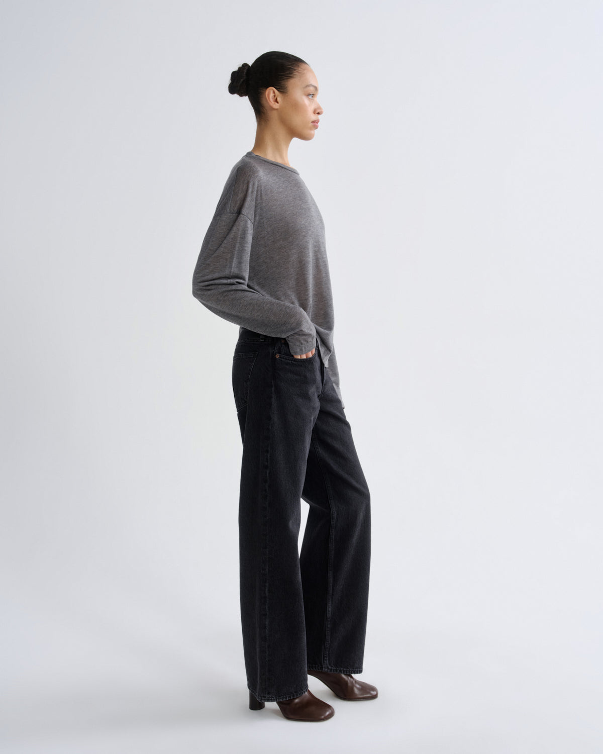 A model standing in profile wearing a gray knit long-sleeve top, dark wide-leg jeans, and brown shoes against a light gray studio background.