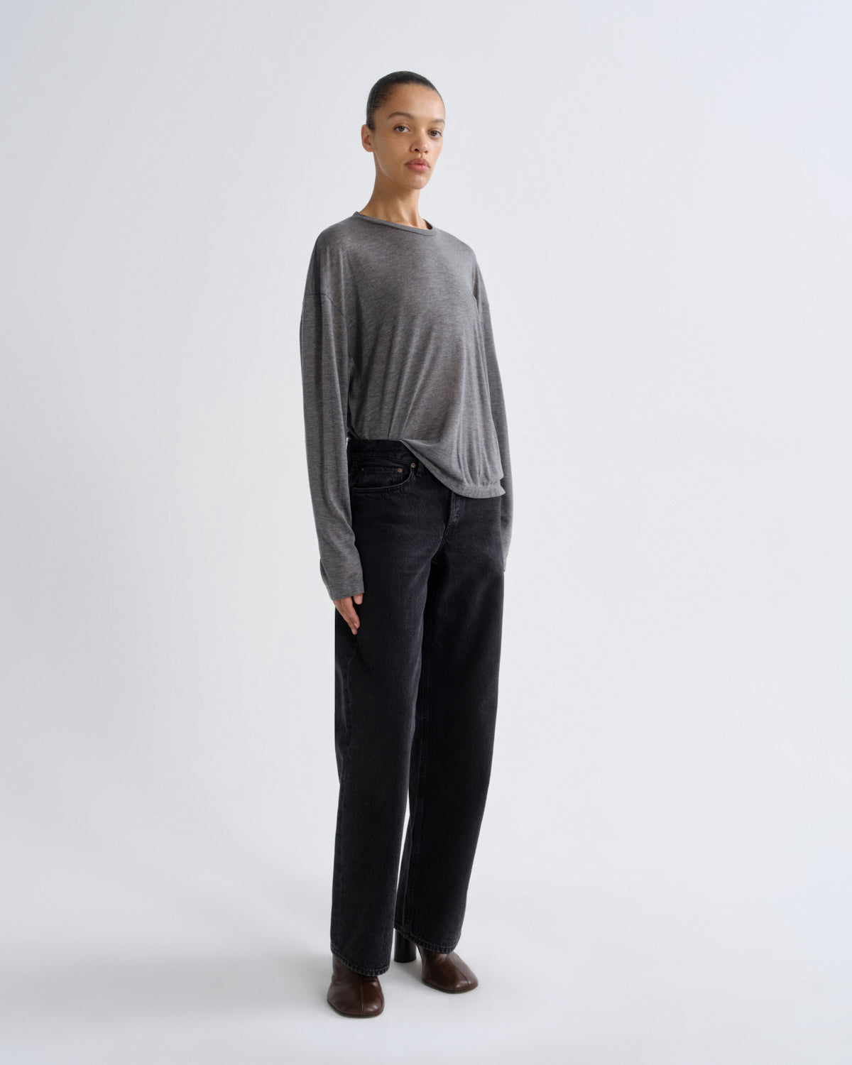 A model stands against a light gray studio background wearing a heather gray long-sleeve knit top, black wide-leg jeans, and brown leather shoes.