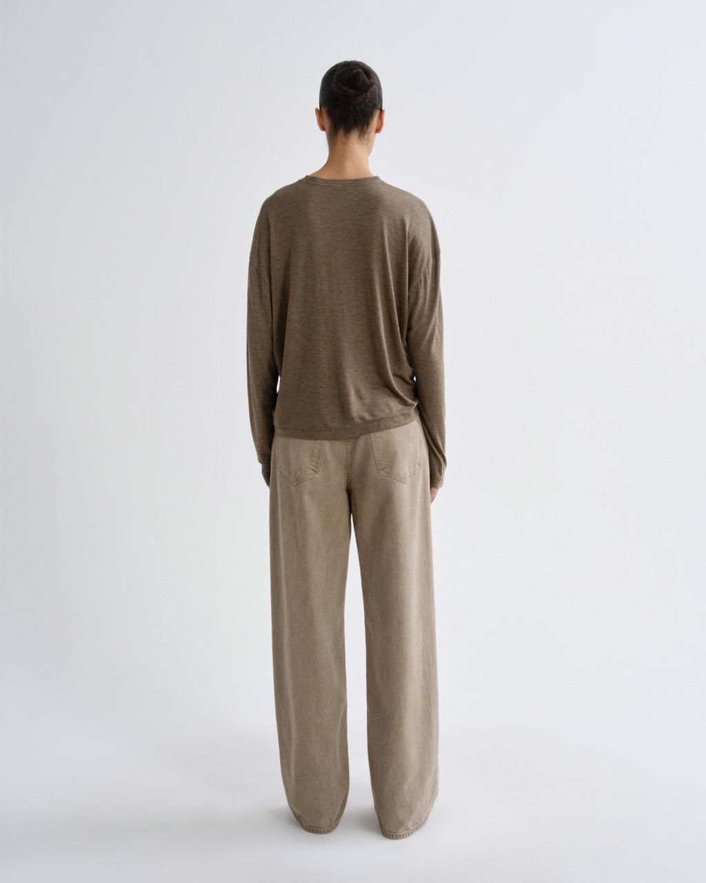 A model viewed from the back wearing a brown long-sleeve crewneck top and beige wide-leg trousers against a light gray studio background.