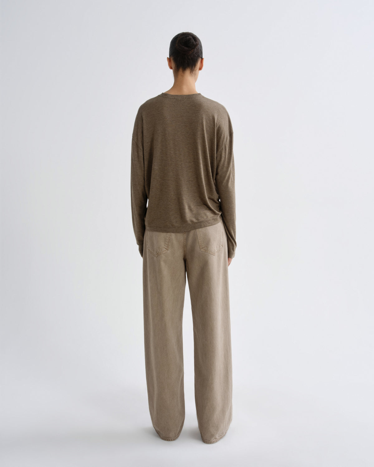 A model viewed from the back wearing a brown long-sleeve crewneck top and beige wide-leg trousers against a light gray studio background.