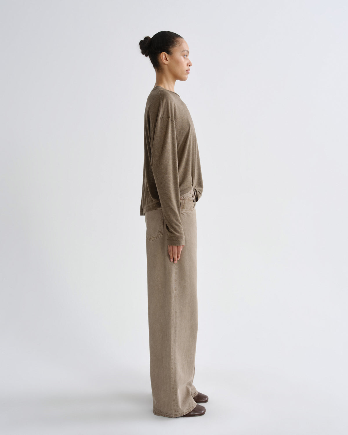 A model in side profile wearing a matching taupe-brown long-sleeve top and wide-leg trousers set, photographed against a clean light gray studio background.