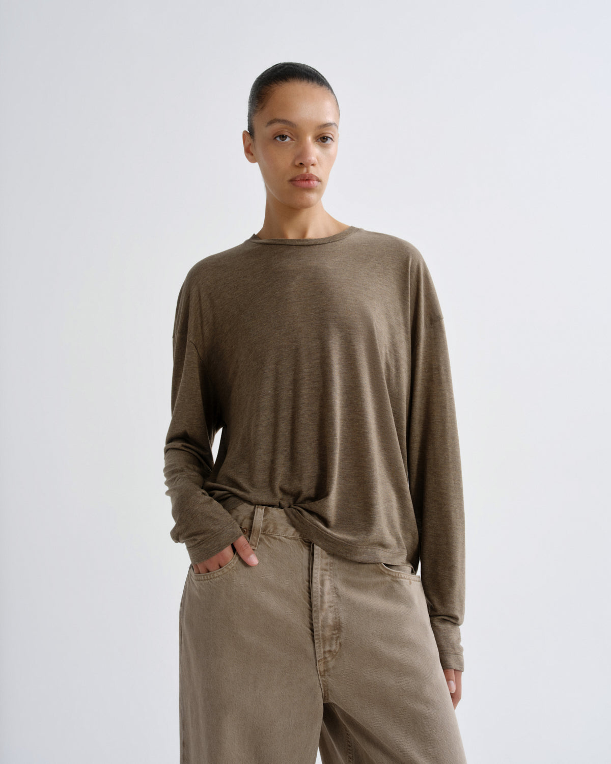 A model wearing a long-sleeve, crew-neck t-shirt in a muted taupe-brown color, paired with light brown corduroy pants, against a plain light gray studio background.