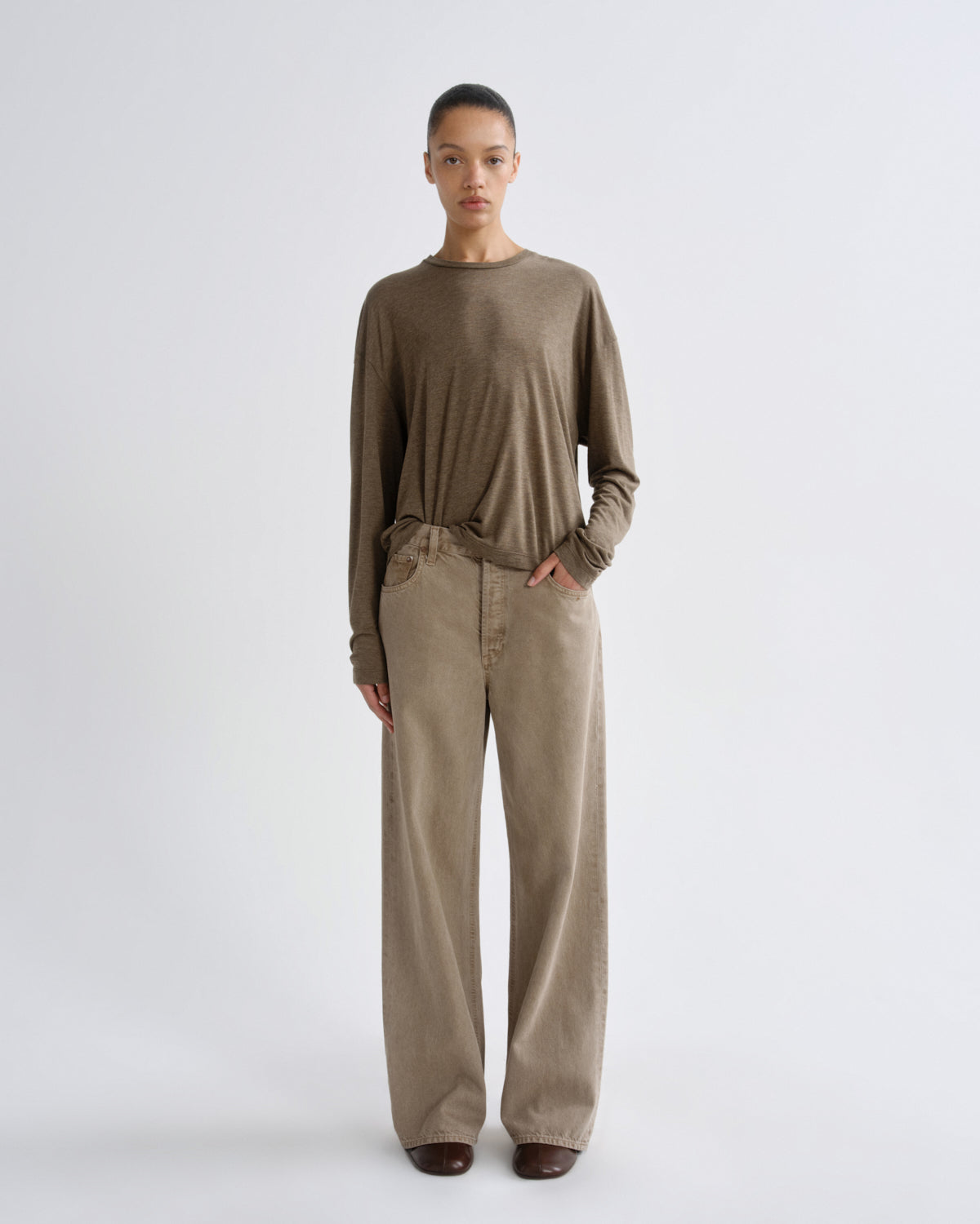 A model stands against a light gray studio background wearing a loose, long-sleeve taupe-brown knit top and matching wide-leg trousers.