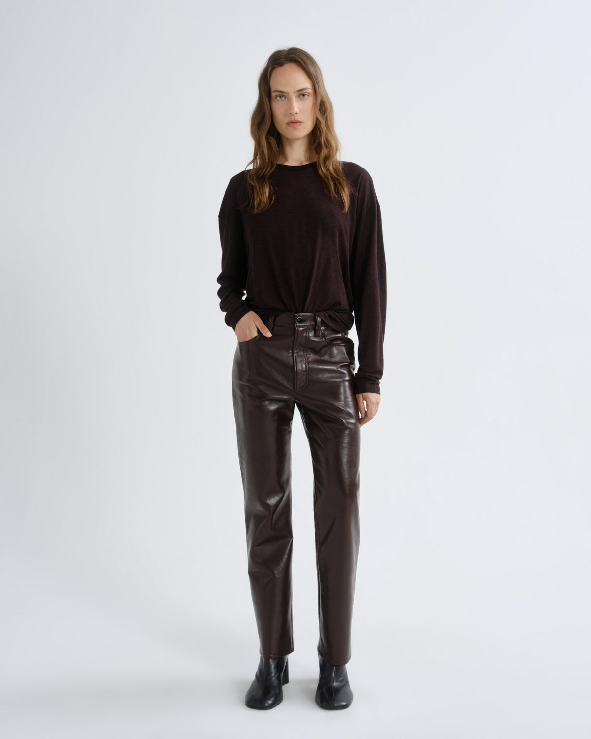 A woman stands against a light gray studio background wearing a dark brown long-sleeve top, dark brown high-waisted pants with a glossy finish, and black ankle boots.
