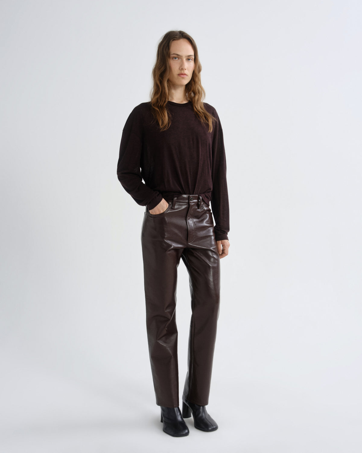 A woman stands against a light gray studio background wearing a dark brown long-sleeve top, shiny dark brown high-waisted pants, and black shoes.