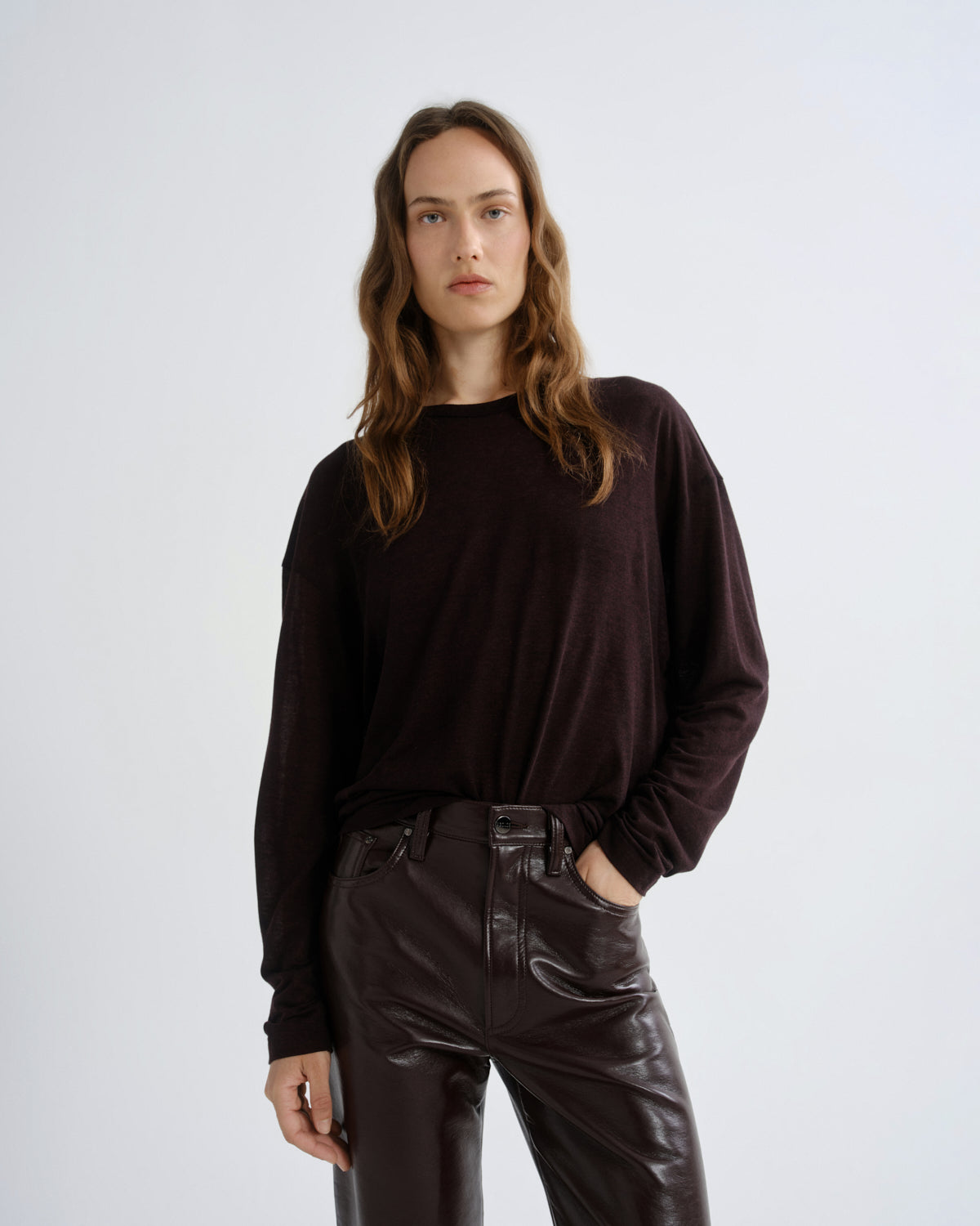 A woman modeling a dark brown long-sleeve crewneck top paired with dark brown faux-leather pants, standing against a light gray studio background.